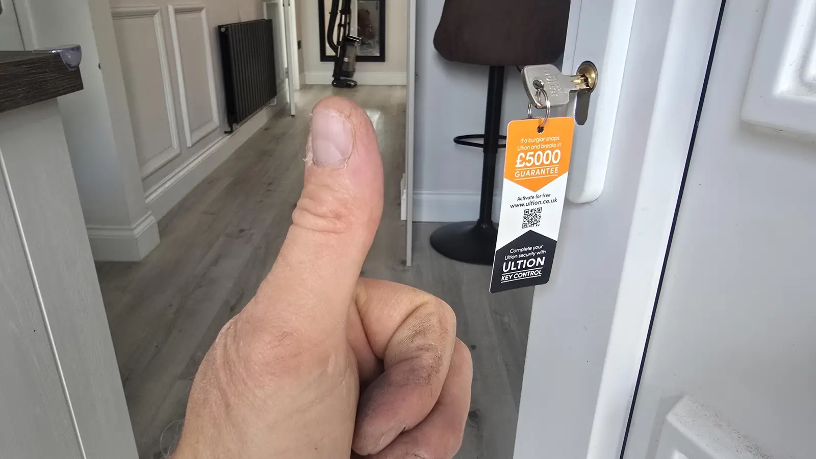 Sean Hamilton giving a thumbs-up beside a brand-new Ultion 3 Star Plus anti-snap euro cylinder he has just fitted to a customer's white front door, the genuine Ultion key and the orange £5000 anti-snap guarantee tag hanging in the lock, the home's hallway visible behind