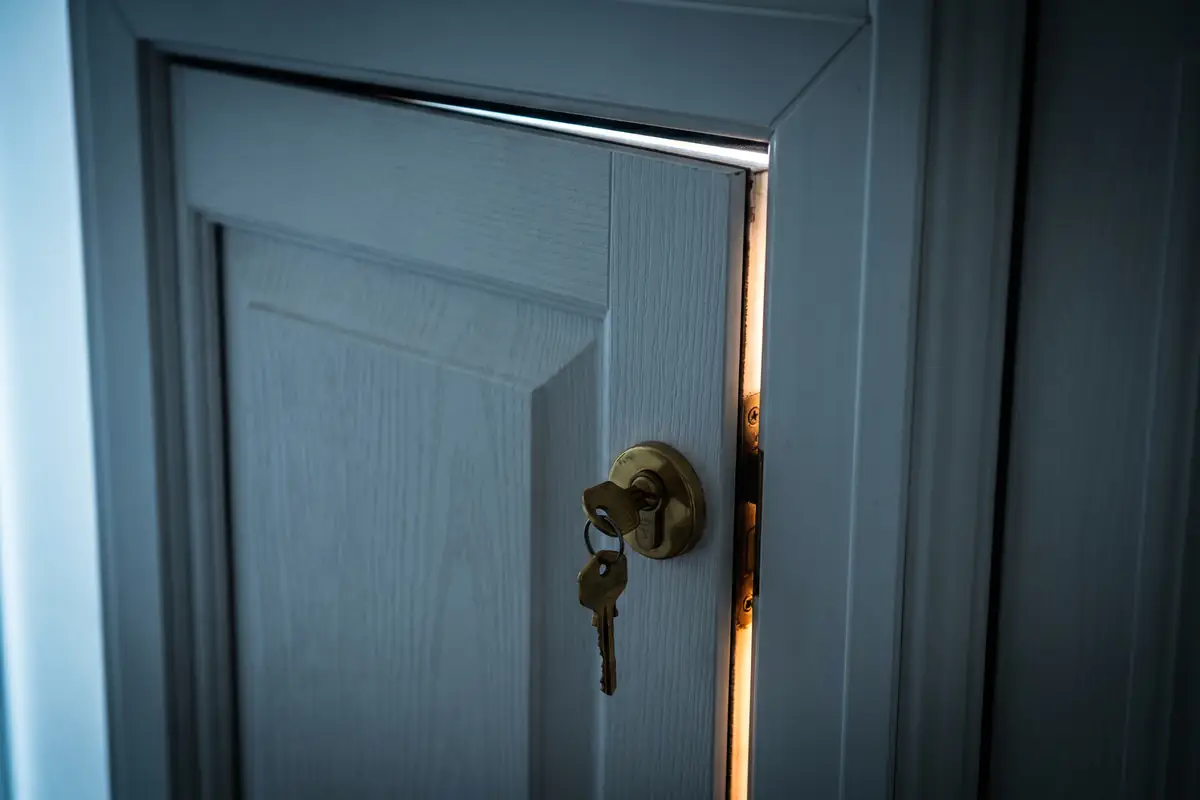 A uPVC composite front door slightly out of square on its hinges, with a small visible gap at the top between door and frame, brass Euro cylinder mid-frame, daylight from the left and warm amber accent from inside the porch